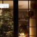 Albero di Natale illuminato visto attraverso una finestra, con luci calde e decorazioni che creano un’atmosfera natalizia in casa.